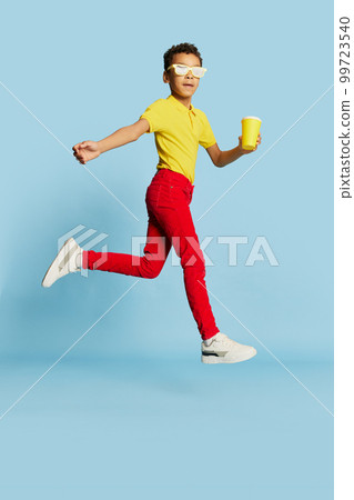 Fashionable little african boy in stylish sunglasses and yellow summer shirt and red trousers posing over blue background. Concept of fashion, happiness, kids emotions Fashionable little african boy in stylish sunglasses and yellow summer shirt and red trousers posing over blue background. Concept of fashion, happiness, kids emotions 99723540