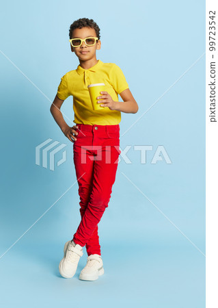 Fashionable little african boy in stylish sunglasses and yellow summer shirt and red trousers posing over blue background. Concept of fashion, happiness, kids emotions 99723542