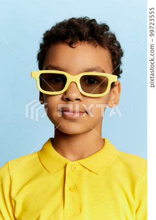 Closeup portrait of little african cute boy in stylish sunglasses and yellow summer shirt looking at camera over blue background. Concept of facial expression, happiness, kids emotions 99723555