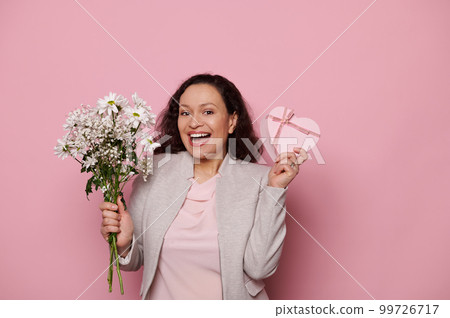 Attractive modern Middle-Eastern woman in elegant light gray jacket, holding a pink pastel heart shaped gift box and a bouquet of flowers, isolated pink background. International Womens Day concept Attractive modern Middle-Eastern woman in elegant light gray jacket, holding a pink pastel heart shaped gift box and a bouquet of flowers, isolated pink background. International Womens Day concept 99726717