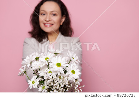 Cute spring bouquet of white chrysanthemums and gypsophila flowers, in hands of a blurred charming multi-ethnic woman, smiling a toothy smile looking at camera, isolated pink background. Copy ad space Cute spring bouquet of white chrysanthemums and gypsophila flowers, in hands of a blurred charming multi-ethnic woman, smiling a toothy smile looking at camera, isolated pink background. Copy ad space 99726725