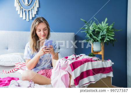 Smiling woman using smartphone to find places where to donate or sale clothes. Decluttering, Sorting clothes and Cleaning Up. Conscious consumer, sustainability. Reuse, second hand, Charity concept Smiling woman using smartphone to find places where to donate or sale clothes. Decluttering, Sorting clothes and Cleaning Up. Conscious consumer, sustainability. Reuse, second hand, Charity concept 99727387