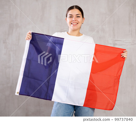 Glad young woman posing and smiling, standing with flag of France against gray wall Glad young woman posing and smiling, standing with flag of France against gray wall 99737840
