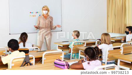 Female teacher in protective mask is giving lecture for primary school students in classroom Female teacher in protective mask is giving lecture for primary school students in classroom 99738213