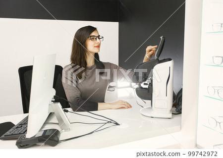 Female optician using laptop at her working place. Woman wearing eyeglasses in ophthalmic shop. Optician checking eyeglasses in special machine Female optician using laptop at her working place. Woman wearing eyeglasses in ophthalmic shop. Optician checking eyeglasses in special machine 99742972