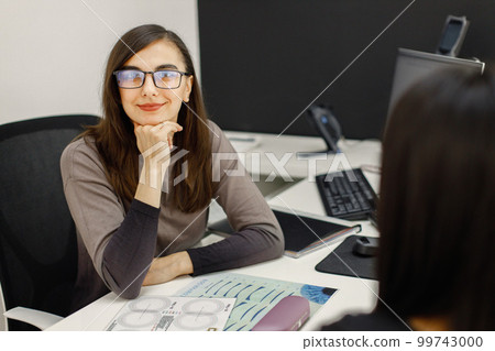 Female optician using laptop and sitting on her working place. Woman wearing eyeglasses in ophthalmic shop. Professional using scheme on the papers to make right choise. 99743000