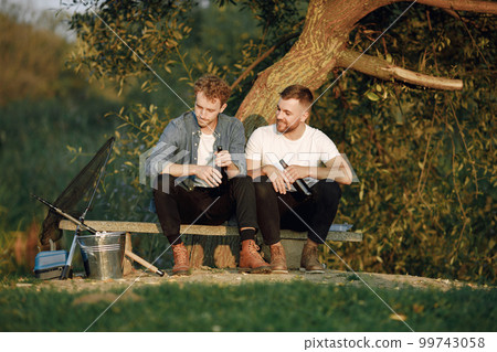 Friends is sitting near a tree and fishing. Two fishermen talking and drinking a beer. One man wearing white t-shirt and other blue shirt. 99743058