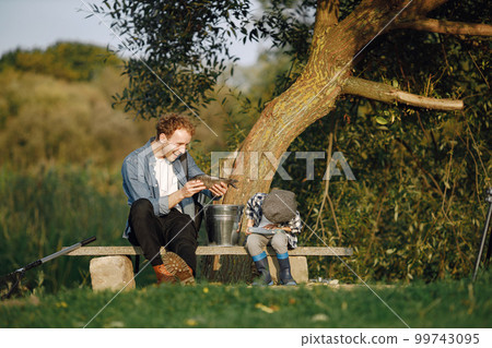 Young dad and his child boy spend time outdoors together. Curly toddler boy wearing a plaid shirt and a hat. Boy sitting near the tree and listening his father. 99743095