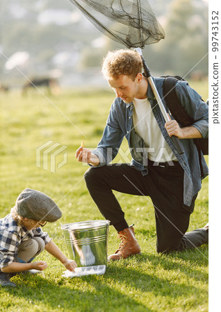 Dad and his child boy spend time outdoors together. Curly toddler boy wearing a plaid shirt and a hat. Boy standing near a bucket and listening his father. Dad and his child boy spend time outdoors together. Curly toddler boy wearing a plaid shirt and a hat. Boy standing near a bucket and listening his father. 99743152