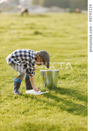 Toddler boy holding a box with a baits for fishing. Boy wearing plaid shirt and a hat. Little boy standing on a grass near bucket. 99743154