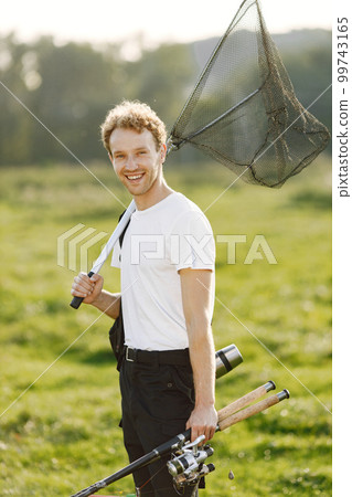 Fisherman is ready to go fishing. Blonde man holding a fishing equipment. Man wearing white t-shirt and black trousers. 99743165