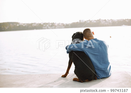 Back view of handsome young male in casual outfit hugging cute dog while sitting near the lake. Boy wearing blue shirt. Dog has a white and black scarf on his neck. Back view of handsome young male in casual outfit hugging cute dog while sitting near the lake. Boy wearing blue shirt. Dog has a white and black scarf on his neck. 99743187