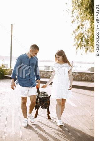 Romantic couple boyfriend and girlfriend walking with rotweiller dog near the lake together. Man and woman looking at each other. Man wearing blue shirt and woman white dress. Romantic couple boyfriend and girlfriend walking with rotweiller dog near the lake together. Man and woman looking at each other. Man wearing blue shirt and woman white dress. 99743201