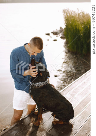 Handsome young male in casual outfit playing with cute dog while standing near the lake. Boy wearing blue shirt and white jeans shorts. Dog has a white and black scarf on his neck. Handsome young male in casual outfit playing with cute dog while standing near the lake. Boy wearing blue shirt and white jeans shorts. Dog has a white and black scarf on his neck. 99743223