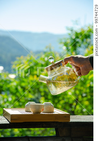 male hand is holding teapot and pouring tea into teacup with tree view background male hand is holding teapot and pouring tea into teacup with tree view background 99744729