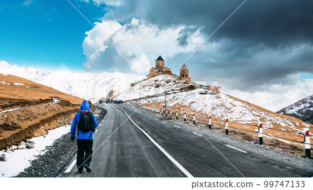 Stepantsminda, Georgia. Man Tourist Backpacker Traveler Photographer Walking To Holy Trinity Church - Tsminda Sameba. Concept Of Pilgrim Or Tourist Traveler. Bright Blue Cloudy Sky Above Church 99747133