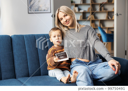 Young pregnant woman and her son sit on sofa at home and boy holds ultrasound scan in hand.They spend time together, enjoy communication feeling kicks of unborn baby 99750491