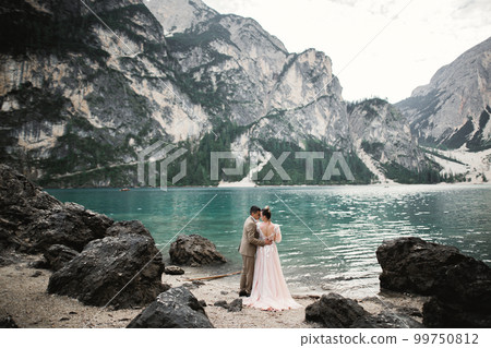Wedding couple on the nature is hugging each other near a beautiful lake in the mountains.. Beautiful model girl in white dress. Man in suit Wedding couple on the nature is hugging each other near a beautiful lake in the mountains.. Beautiful model girl in white dress. Man in suit 99750812