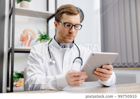 Male Caucasian doctor in white medical uniform filling patient medical history or anamnesis on tablet in office. Doctor consulting client online on tablet in modern clinic, healthcare concept Male Caucasian doctor in white medical uniform filling patient medical history or anamnesis on tablet in office. Doctor consulting client online on tablet in modern clinic, healthcare concept 99750910