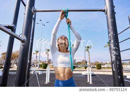 Active lifestyle. Slim young woman training pull ups with resistance band in outdoors gym at sunny 99755124