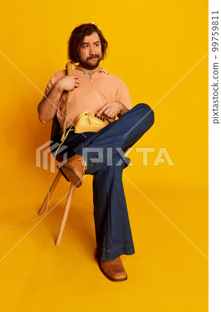 Emotional bearded man, hippie in vintage style flared jeans sitting and talking on retro phone over yellow background. Concept of fashion, emotions, american culture 99759811
