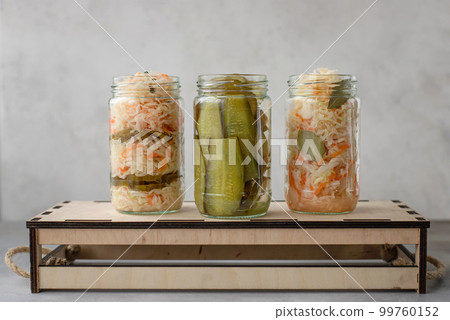 various fermented cabbage and cucumbers in jars on a wooden box. 99760152