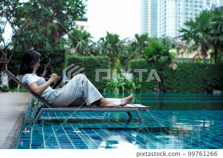 Portrait Asian woman sits on the bench in the swimming pool and writes on the screen by the Bluetooth pen with defocused trees in background 99761266