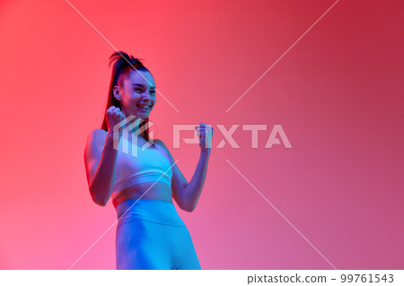 Emotions of win. Portrait of happy young female athlete in uniform posing over pink studio background in neon light. Concept of sport. health and strength Emotions of win. Portrait of happy young female athlete in uniform posing over pink studio background in neon light. Concept of sport. health and strength 99761543