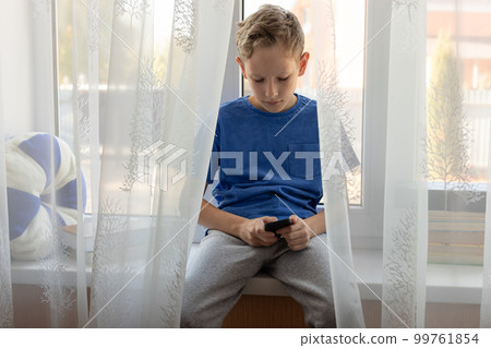 child boy typing message on smartphone while sitting on windowsill at home. 99761854