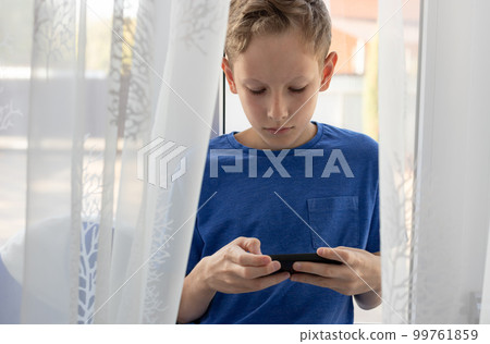 child boy uses mobile phone while sitting on the windowsill at home. 99761859