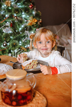 A girl with blond hair in pajamas eats a pie at a table near the Christmas tree 99762152