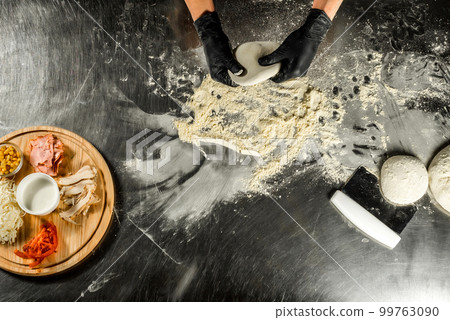 Cooking pizza on a steel table; dough, flour, pizza topping, sauce and chef`s hands in black gloves on the table 99763090