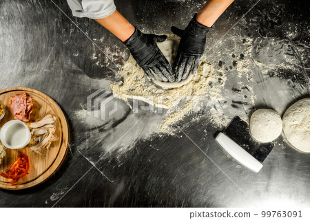 Cooking pizza on a steel table; dough, flour, pizza topping, sauce and chef`s hands in black gloves on the table Cooking pizza on a steel table; dough, flour, pizza topping, sauce and chef`s hands in black gloves on the table 99763091