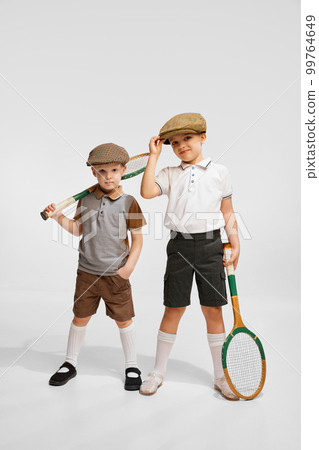 Two little boys, children in retro clothes posing with tennis rackets against grey studio background. Concept of childhood 99764649