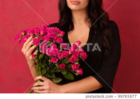 A young woman in a black dressing gown with a deep neckline holds a bouquet of pink roses, on a pink background, close-up 99765877