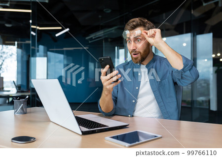 A young man works in the office at a table using a laptop. He holds the phone in his hands, looks at the screen happy and shocked, raised his glasses on his face in surprise. Got good news. A young man works in the office at a table using a laptop. He holds the phone in his hands, looks at the screen happy and shocked, raised his glasses on his face in surprise. Got good news. 99766110