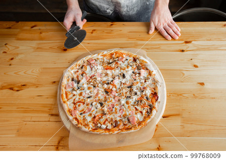 Closeup hand of chef baker in uniform blue apron cutting pizza at kitchen Closeup hand of chef baker in uniform blue apron cutting pizza at kitchen 99768009