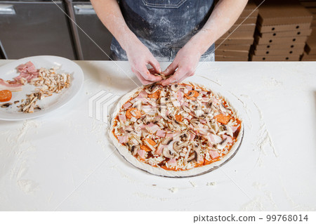 Cooking pizza. Lays out the cheese and salami filling, ingredients on the dough preform. Closeup hand of chef baker in uniform white apron cook at kitchen Cooking pizza. Lays out the cheese and salami filling, ingredients on the dough preform. Closeup hand of chef baker in uniform white apron cook at kitchen 99768014