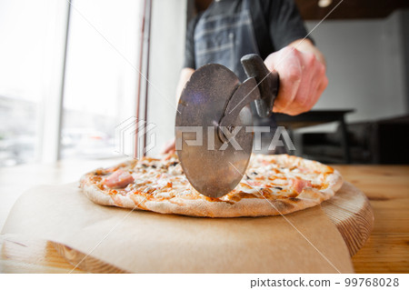 Closeup hand of chef baker in uniform blue apron cutting pizza at kitchen 99768028