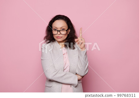 Attractive confident multi-ethnic woman with beautiful curly brown hair, wearing stylish spectacles and formal suit, points index finger up at copy space, looking at camera on isolated pink background 99768311