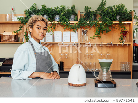 Young woman in apron working as a barista standing at the counter with a kettle, filter and scales Young woman in apron working as a barista standing at the counter with a kettle, filter and scales 99771541