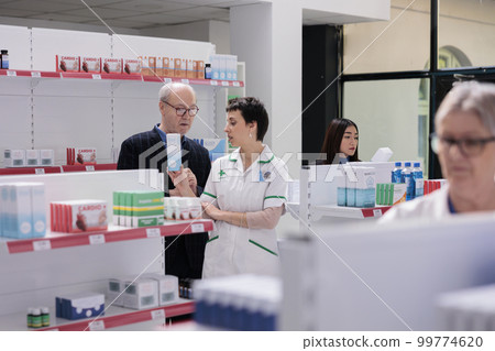 Young woman pharmacist reading usage instruction on sunblock cream box to senior customer, standing in medical retail store aisle. Buyer consulting with pharmacy worker about dermatologic product Young woman pharmacist reading usage instruction on sunblock cream box to senior customer, standing in medical retail store aisle. Buyer consulting with pharmacy worker about dermatologic product 99774620