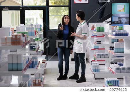Drugstore buyer asking pharmacist for dietary supplements usage advice. Pharmaceutical assistant and young asian woman standing in pharmacy aisle, discussing multivitamin dosage instruction Drugstore buyer asking pharmacist for dietary supplements usage advice. Pharmaceutical assistant and young asian woman standing in pharmacy aisle, discussing multivitamin dosage instruction 99774625