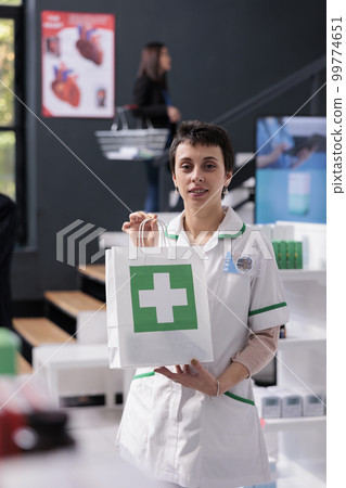 Pharmacist woman selling medicaments and holding paper shopping bag portrait. Pharmaceutical consultant helping choosing pills, giving madications purchase and looking at camera Pharmacist woman selling medicaments and holding paper shopping bag portrait. Pharmaceutical consultant helping choosing pills, giving madications purchase and looking at camera 99774651