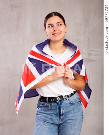 Joyous young woman holding Britain flag against unicoloured background Joyous young woman holding Britain flag against unicoloured background 99775724