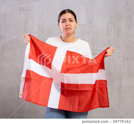Upset young woman holding flag of Denmark against gray wall indoors Upset young woman holding flag of Denmark against gray wall indoors 99776162