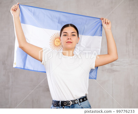 Excited young woman holding state flag of Argentina against gray wall background indoors 99776233