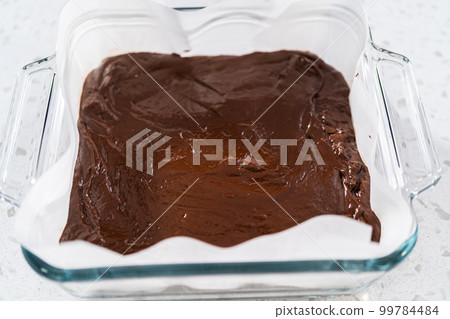 Pouring fudge mixture into the baking pan lined with parchment paper to prepare chocolate fudge with peanut butter swirl. 99784484