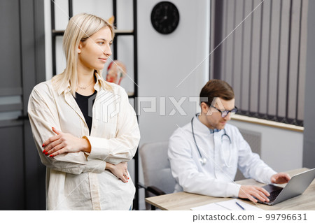 A young attractive doctor is having a consultation in his office. The doctor is making notes on the laptop, and the patient is standing by the table, smiling 99796631