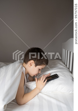 A 5 year old boy plays games on his phone while lying in bed. Gadgets in bed before bedtime in a child A 5 year old boy plays games on his phone while lying in bed. Gadgets in bed before bedtime in a child 99797016